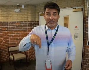 A man wearing a light-colored button-up shirt and lanyard stands indoors near brick walls, pointing downward with a surprised facial expression. A chair and an open door are visible in the background. | High 5 Plumbing, Heating & Cooling