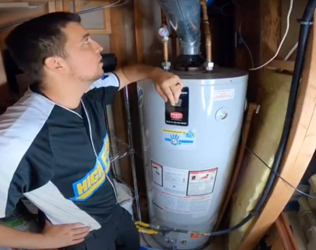 A man in a black and white shirt stands next to a water heater in a utility room, resting his arm on top of the unit and looking up at the pipes and connections above it. | High 5 Plumbing, Heating & Cooling
