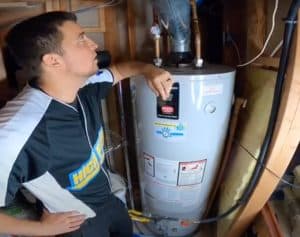 A man in a black and white shirt stands next to a water heater in a utility room, resting his arm on top of the unit and looking up at the pipes and connections above it. | High 5 Plumbing, Heating & Cooling