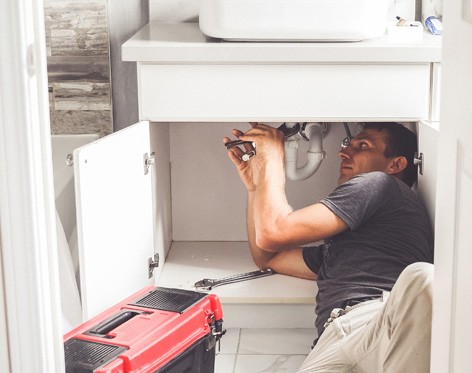 A person lying on the floor works under a bathroom sink with a wrench. The cabinet doors are open, and a red toolbox is nearby on the tiled floor. | High 5 Plumbing, Heating & Cooling