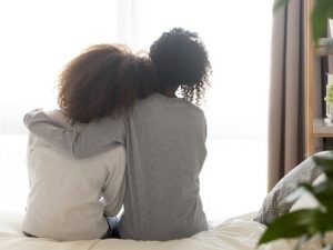 Two people with curly hair sit close together on a bed, seen from behind. One person has their arm around the other. Soft natural light comes through a window in the background. | High 5 Plumbing, Heating & Cooling