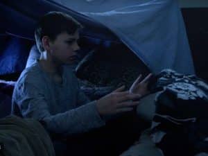 A boy wearing a gray long-sleeve shirt sits inside a dimly lit blanket fort, reaching forward with both hands toward a pile of pillows and blankets. | High 5 Plumbing, Heating & Cooling