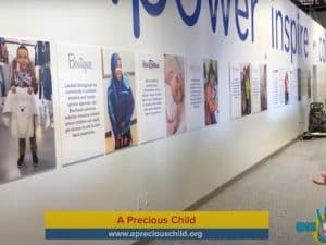 A wall display features large photos and informational posters about community services. The wall has words like “power” and “inspire.” The bottom of the image shows a “A Precious Child” banner with a website link. | High 5 Plumbing, Heating & Cooling