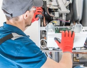 A technician wearing a blue shirt, gray cap, and red gloves is inspecting and repairing the internal components of a gas boiler or furnace, with various wires and circuit boards visible. | High 5 Plumbing, Heating & Cooling