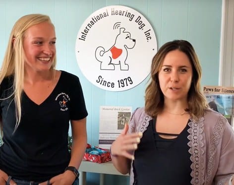 Two women stand indoors in front of a wall sign reading "International Hearing Dog, Inc. Since 1979." One woman is smiling while the other is speaking and gesturing with her hand. Pamphlets and a gift box are visible behind them. | High 5 Plumbing, Heating & Cooling