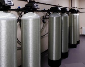Five large industrial water filtration tanks with digital control panels are lined up against a wall, interconnected by pipes and hoses in a clean, tiled utility room. | High 5 Plumbing, Heating & Cooling