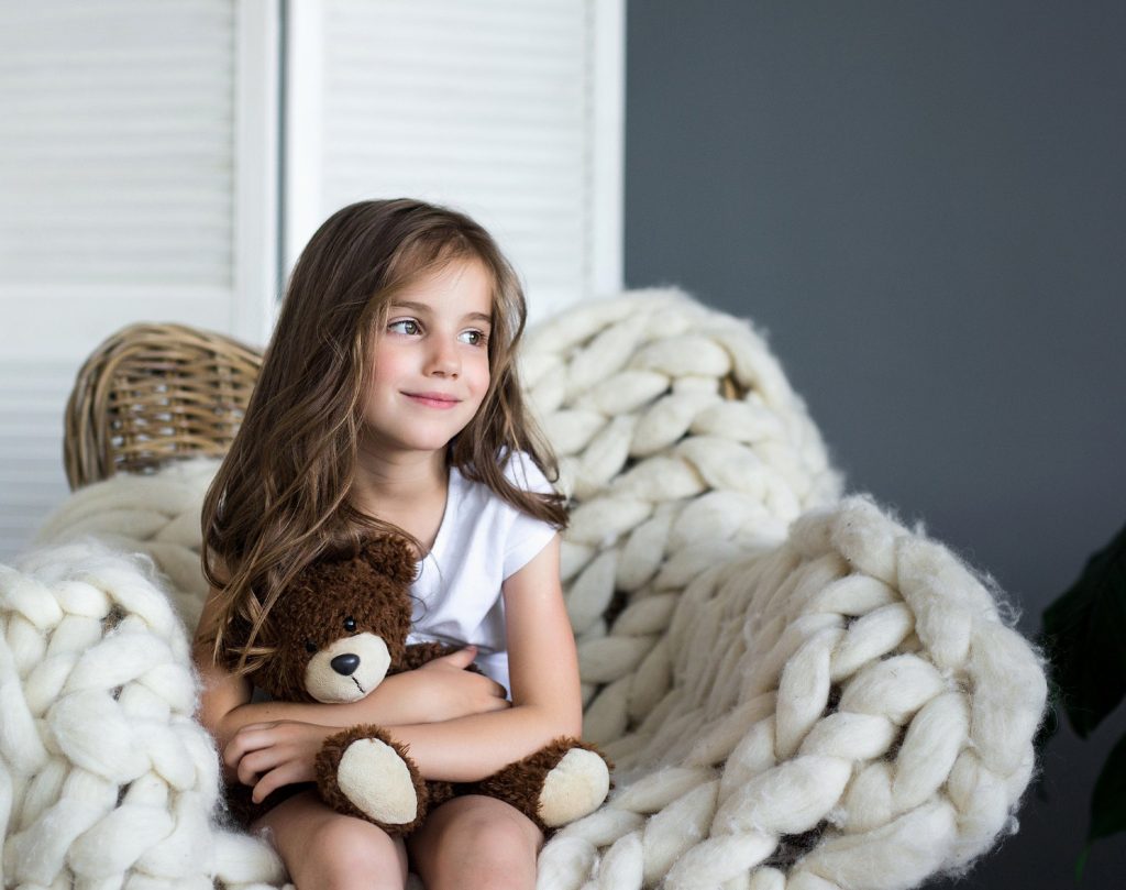 A young girl with long brown hair sits on a chunky white knitted chair, holding a brown teddy bear and smiling slightly. The background includes a wicker basket and a gray wall. | High 5 Plumbing, Heating & Cooling