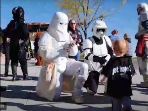 Two people dressed as Star Wars stormtroopers kneel on a paved area outdoors, interacting with a young child wearing a black "got hope?" shirt; several adults and trees are visible in the background. | High 5 Plumbing, Heating & Cooling