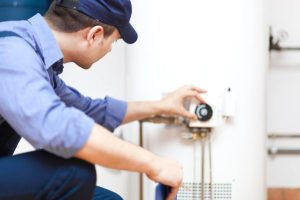 A person wearing a blue uniform and cap is performing maintenance by adjusting the control panel on a water heater in an indoor basement setting. | High 5 Plumbing, Heating & Cooling