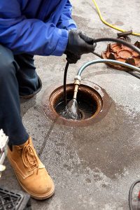 A person wearing a blue jacket, black gloves, and tan work boots is performing sewer repair, holding a hose and inserting it into an open drain or manhole on a concrete surface. Water and equipment are visible around the opening. | High 5 Plumbing, Heating & Cooling