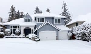 A two-story suburban house with snow covering the roof, driveway, and yard. Evergreen trees and bushes are blanketed in snow, while the overcast sky hints at a recent snowfall—perfect weather to check and winterize plumbing systems. | High 5 Plumbing, Heating & Cooling