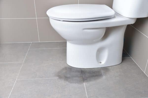 A white toilet sits against a tiled wall on a gray tiled floor. A visible wet stain near the base suggests potential toilet maintenance is needed to prevent further issues. | High 5 Plumbing, Heating & Cooling