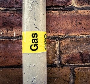 A gray pipe with a yellow "Gas" label runs vertically in front of a brick wall, showing water droplets on its surface—a sign that gas line repair may be necessary. | High 5 Plumbing, Heating & Cooling