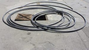A large metal manhole cover is partially open on a paved surface, with several long black cables coiled loosely around it—part of important work to prevent sewage backups in the area. | High 5 Plumbing, Heating & Cooling