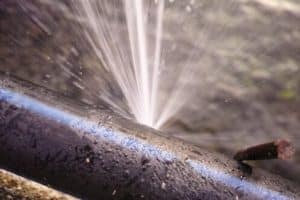 A close-up of a black pipe with a blue stripe, spraying water forcefully from a pipes burst. Water droplets are visible on the pipe, and a rusty metal rod is nearby. | High 5 Plumbing, Heating & Cooling