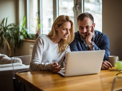 A woman and a man sit at a wooden table, looking at a laptop screen together. The man holds a mug and an apple is on the table as they research the benefits of installing a yard hydrant. Large windows and a plant are in the background. | High 5 Plumbing, Heating & Cooling