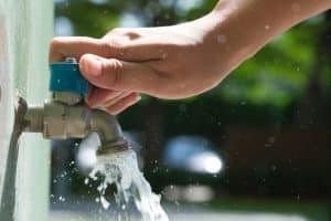 A person’s hand is turning on a blue-handled outdoor water faucet, with water flowing out of the tap and droplets splashing—highlighting reliable access, much like gas plumbing during a power outage. The background is blurred greenery and light. | High 5 Plumbing, Heating & Cooling