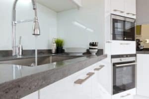 Modern kitchen with white cabinets, a built-in oven and microwave, a stainless steel sink with a high-arc faucet showing hard water signs, and stacked dishes and cups on the countertop near a green potted plant. | High 5 Plumbing, Heating & Cooling