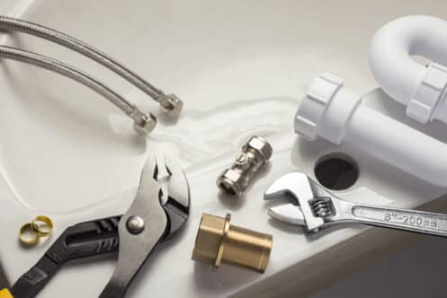 Plumbing tools and materials for bathtub repair are laid out on a white surface next to a sink. Items include a wrench, pliers, pipe fittings, valves, braided hoses, a plastic pipe, and brass components—everything needed to fix bathtub cracks. | High 5 Plumbing, Heating & Cooling