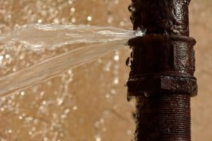 Water sprays from a crack in a rusty metal pipe, signaling a pipe burst. The blurred, water-stained background highlights the need for emergency plumbing. | High 5 Plumbing, Heating & Cooling