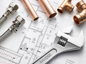 Plumbing tools, copper pipes, flexible hoses, and fittings are arranged on top of architectural blueprints—perfect for a home repiping project. A large adjustable wrench is also visible in the foreground. | High 5 Plumbing, Heating & Cooling