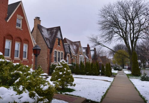 A row of brick and stone houses with steep roofs sits beside a sidewalk. Snow covers the lawns, bushes, and trees, signaling winter—an important time to protect outdoor faucets. The sidewalk and street remain mostly clear under the overcast sky. | High 5 Plumbing, Heating & Cooling