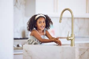 A young girl with curly hair and a headband is smiling while washing her hands under a running faucet with water filtration at a marble kitchen sink. The background shows a modern kitchen with white cabinets. | High 5 Plumbing, Heating & Cooling