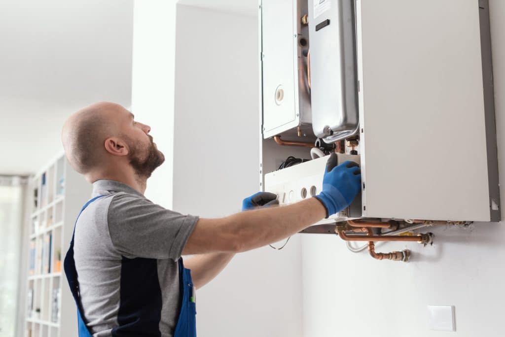 A man wearing blue gloves and overalls is servicing a wall-mounted boiler unit, commonly found in residential water heating systems. He is looking up at the boiler, with his hands adjusting the front panel in a home setting. | High 5 Plumbing, Heating & Cooling