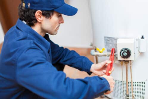 A person wearing a blue uniform and cap uses a red pipe wrench to adjust a component on a white water heater, showcasing emergency plumbing skills. Various pipes and a control panel are visible on the heater. | High 5 Plumbing, Heating & Cooling