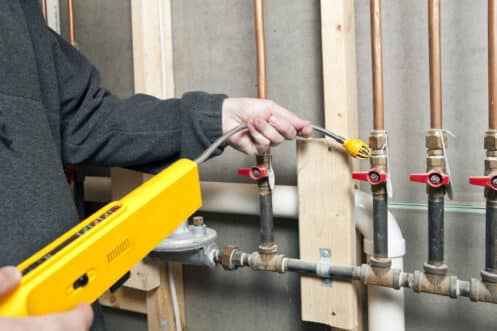 A person uses a handheld electronic gas leak detector to inspect copper gas pipes for gas line damage in a utility area with unfinished wooden studs and exposed plumbing. | High 5 Plumbing, Heating & Cooling