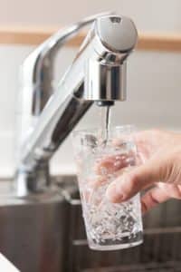 A hand holds a clear glass under a kitchen faucet as water flows in, filtered by a whole-house water filter. The background reveals part of a metallic sink and countertop. | High 5 Plumbing, Heating & Cooling