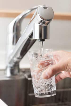 A hand holds a clear glass under a kitchen faucet as water flows in, filtered by a whole-house water filter. The background reveals part of a metallic sink and countertop. | High 5 Plumbing, Heating & Cooling