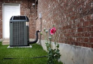 An outdoor air conditioning unit stands on a concrete pad beside a brick house. Poppy flowers bloom in the summer grass nearby, and a black hose lies on the ground. A white door is visible in the background, offering ways to reduce AC costs. | High 5 Plumbing, Heating & Cooling
