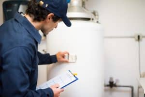A worker in a blue uniform and cap inspects a water heater, holding a clipboard and adjusting the thermostat—checking for signs that an upgrade may be needed in the utility room. | High 5 Plumbing, Heating & Cooling
