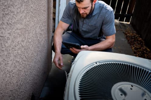 A man in a gray shirt crouches next to an outdoor air conditioning unit, using a tablet to assist with air conditioner troubleshooting as he inspects the equipment beside a building and a wooden fence. | High 5 Plumbing, Heating & Cooling