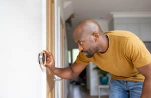 A man wearing a mustard yellow t-shirt adjusts a round thermostat on a white wall in a modern home, preparing his HVAC system for the fall season. He is leaning forward and focusing on the device. | High 5 Plumbing, Heating & Cooling
