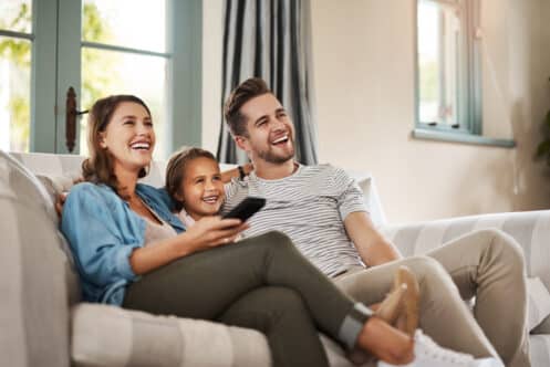 A woman, a man, and a young girl sit together on a couch in a living room, smiling and looking at something off-camera. The woman holds a remote control in their smart home, filled with natural light from energy-efficient windows behind them. | High 5 Plumbing, Heating & Cooling