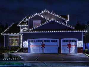 A two-story house at night features Outdoor Holiday Lighting with white Christmas lights along the roofline and driveway. The garage doors have red ribbon designs, and a lit Christmas tree is seen through a front window. | High 5 Plumbing, Heating & Cooling