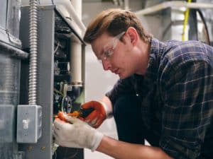 A person wearing safety glasses, gloves, and a plaid shirt uses a screwdriver to work on electrical components inside an open metal panel, preparing for a winter furnace tune-up in an industrial or mechanical setting. | High 5 Plumbing, Heating & Cooling