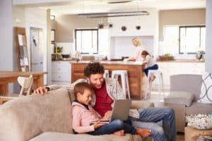A man and a boy sit on a couch looking at a laptop, both smiling—perhaps researching installing a backup generator for winter. In the background, a woman and girl are at the kitchen island in their bright, modern open-plan living space. | High 5 Plumbing, Heating & Cooling