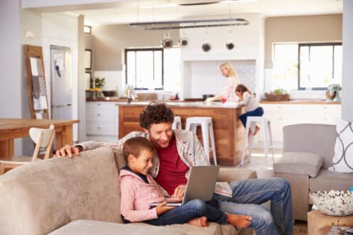 A man and a boy sit on a couch looking at a laptop, both smiling—perhaps researching installing a backup generator for winter. In the background, a woman and girl are at the kitchen island in their bright, modern open-plan living space. | High 5 Plumbing, Heating & Cooling