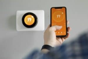 A person holds a smartphone displaying a thermostat app set to 77 degrees, while a smart thermostat on the wall behind them also shows 77 degrees—perfect for year-round comfort. | High 5 Plumbing, Heating & Cooling