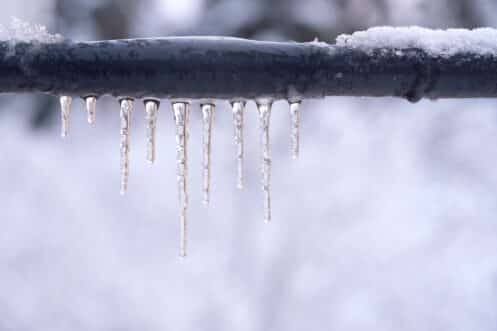 A row of icicles hangs from a snow-covered black metal pipe, with a blurred snowy background—a reminder of the importance of safely thawing pipes to prevent plumbing disasters during harsh winter weather. | High 5 Plumbing, Heating & Cooling
