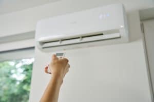 A person points a remote control at a wall-mounted air conditioner, showcasing ductless cooling set to 21 degrees Celsius. A window with greenery outside is partially visible on the left side of the image. | High 5 Plumbing, Heating & Cooling