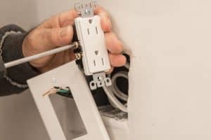 A person is converting a wall fixture by installing a white outlet using a screwdriver. The outlet cover and wires are visible, and the unfinished wall shows where the light switch or outlet is being fitted. | High 5 Plumbing, Heating & Cooling