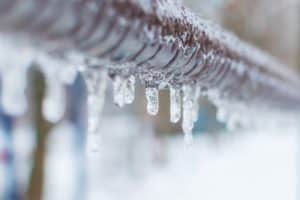 Close-up of icicles hanging from a metal fence or railing, highlighting the cold winter weather and the risk of frozen pipe problems, as ice formations glisten against a snowy, blurred background. Frozen pipes rarely thaw on their own. | High 5 Plumbing, Heating & Cooling