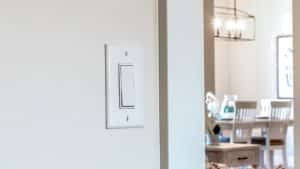 A white dimmer switch on a beige wall in the foreground; in the background, a dining area with a table, chairs, and a chandelier is visible. | High 5 Plumbing, Heating & Cooling