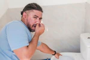 A man in a blue shirt holds his nose and points at a toilet, appearing to react to sewage odors in a bathroom with light-colored tiled walls, possibly hinting at common causes of bad smells in the home. | High 5 Plumbing, Heating & Cooling