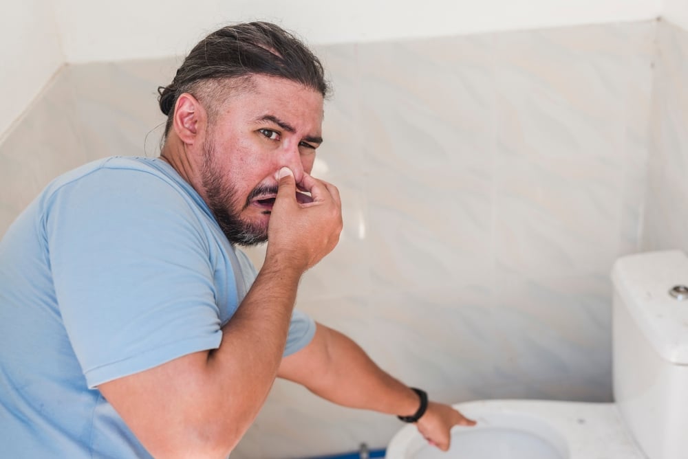 A man in a blue shirt holds his nose and points at a toilet, appearing to react to sewage odors in a bathroom with light-colored tiled walls, possibly hinting at common causes of bad smells in the home. | High 5 Plumbing, Heating & Cooling