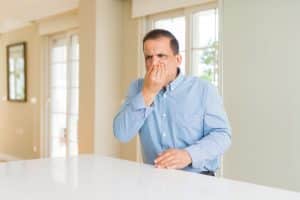 A man in a blue button-up shirt sits at a white table indoors, holding his nose with a displeased expression, as if reacting to a sewer gas smell. Large windows and light-colored walls are visible in the background. | High 5 Plumbing, Heating & Cooling
